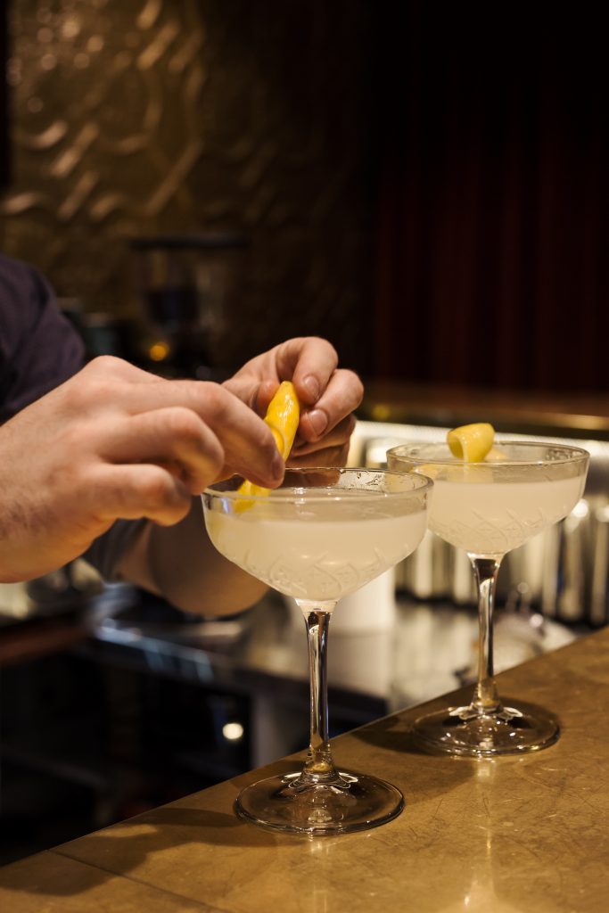 Two drinks in martini glasses on a gold bar, with a bar tender putting the finishing touches on the second drink.